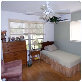 Small bedroom with a single bed, wooden dresser and nightstand, an armchair, ceiling fan, and windows with blinds.