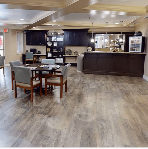 A spacious common area in a senior living facility featuring a round table with four cushioned chairs, a dark wood reception or serving counter with pendant lights above, and built-in dark wood cabinetry along the back wall with decorative items and a small computer workstation.