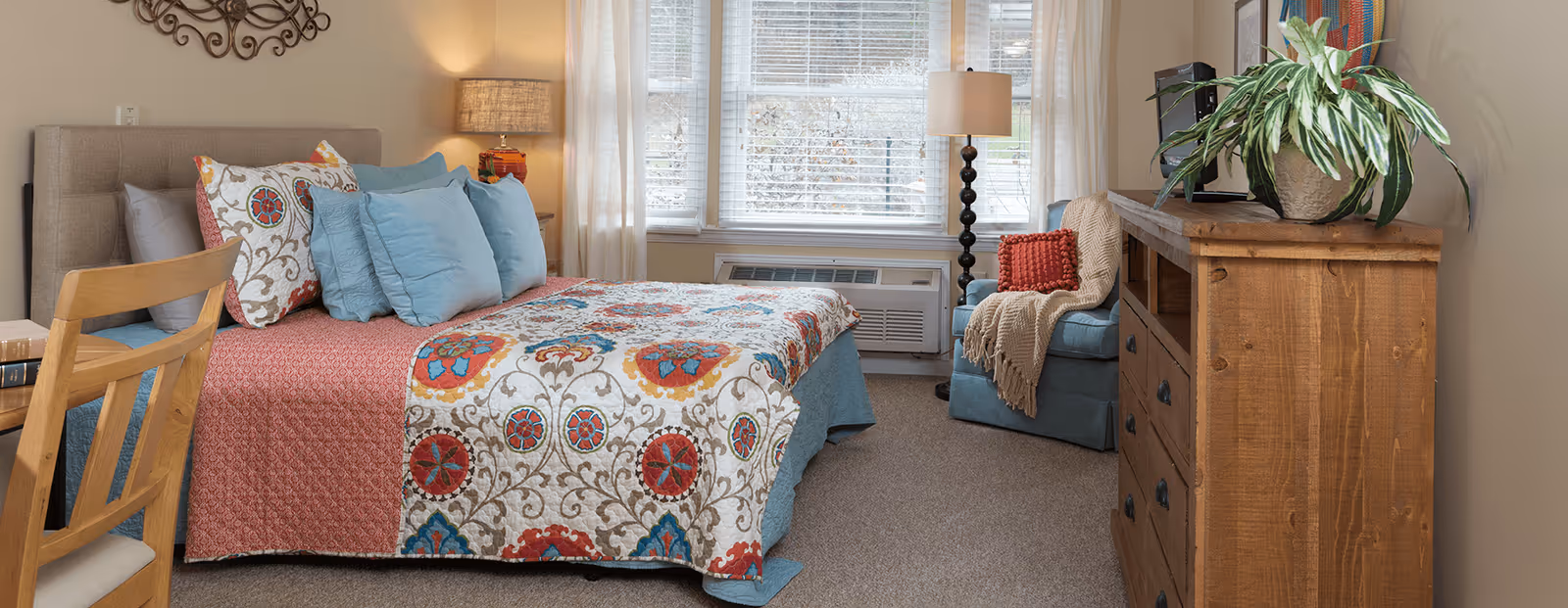 Sunlit furnished bedroom with a patterned bedspread and pillows, wooden dresser, armchair, and a window with blinds.