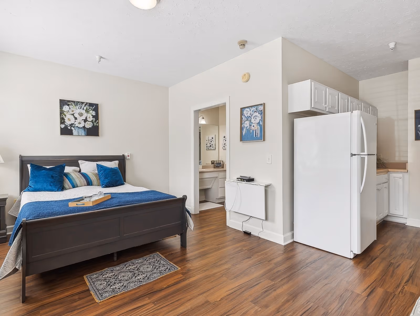 A senior living facility room featuring a dark wooden bed with blue and striped pillows and a blue throw blanket. The room has light-colored walls with floral paintings, wood flooring, and a white refrigerator next to a small kitchen area. A bathroom is visible through an open door.