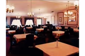 Spacious dining room with multiple tables covered in white tablecloths, upholstered chairs, chandeliers, and draped windows.