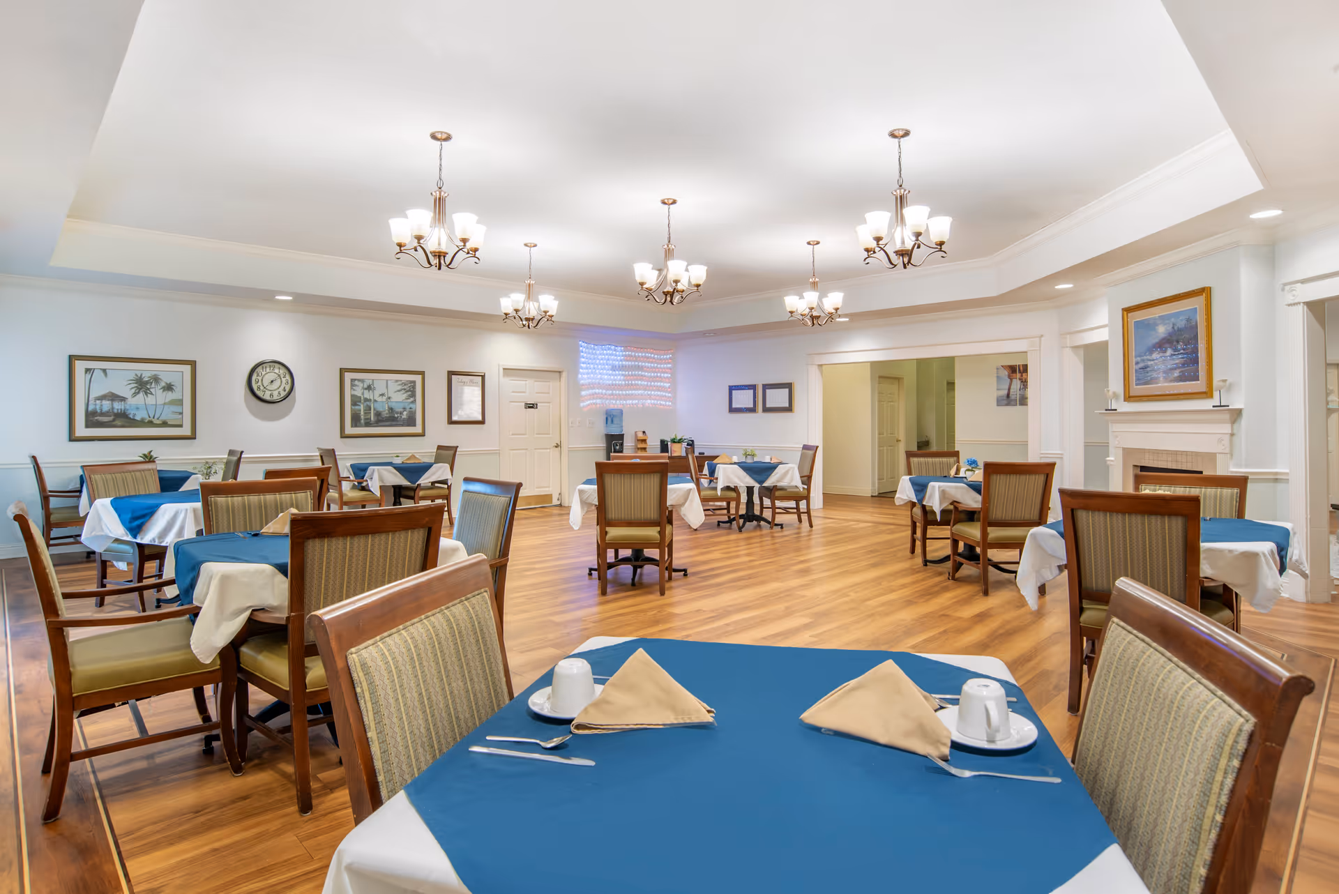 Bright dining room with multiple tables set with blue tablecloths and place settings, wooden chairs, chandeliers, and hardwood floors.