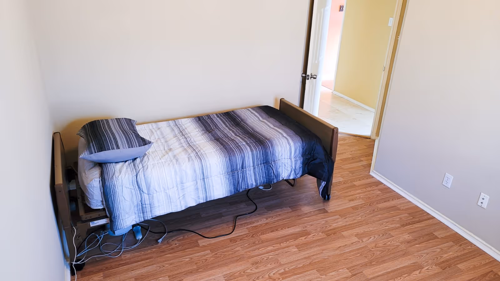 A small bedroom with a single bed against the wall. The bed has a striped blue and white comforter and a matching pillow. The floor is wooden, and there is an open door leading to a hallway with a yellow wall and tiled floor.