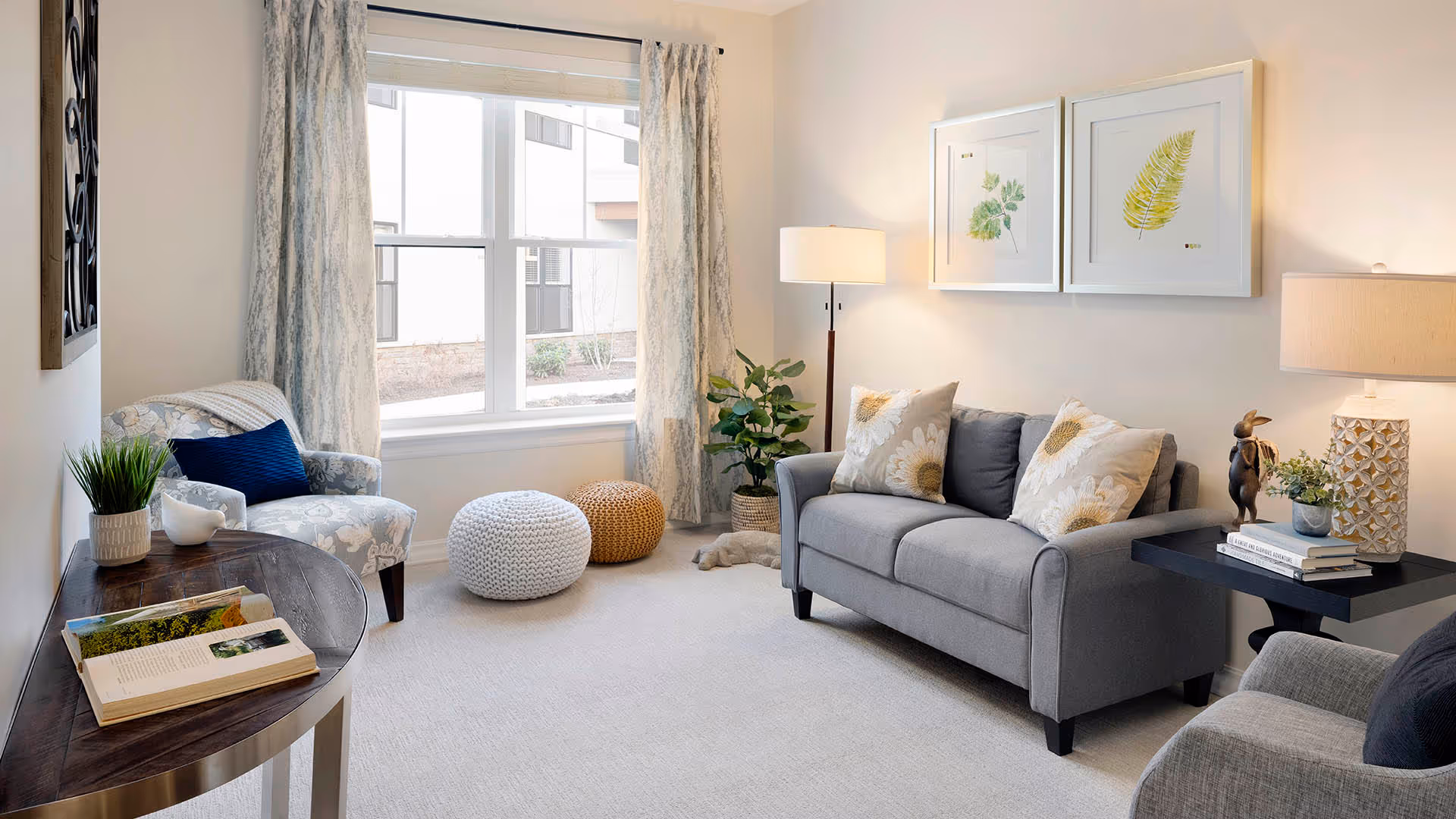Bright, tastefully decorated living room with a gray sofa and patterned pillows, an armchair, poufs, side tables with lamps, and a large window.
