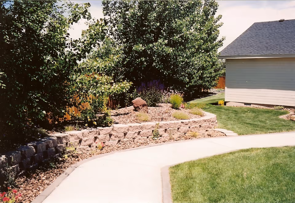 Curved concrete pathway bordered by a low stone retaining wall with various plants and shrubs, leading past a grassy lawn area towards a light gray building with a dark shingled roof under a partly cloudy sky.