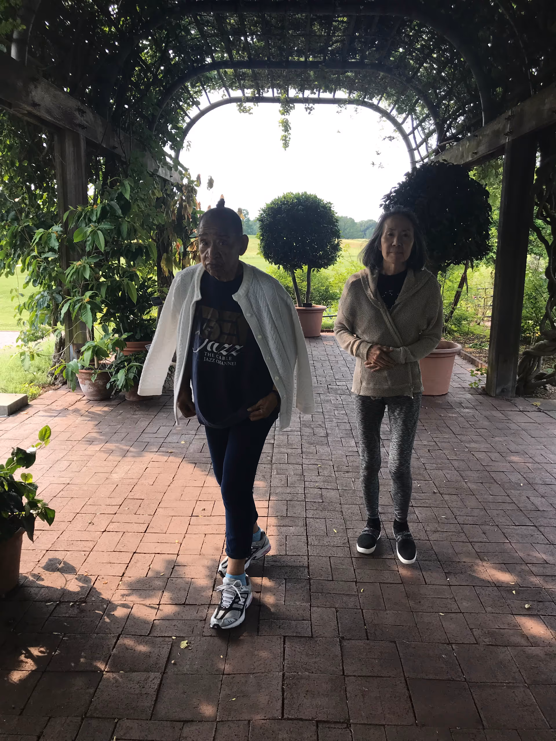 Two elderly women walking under a wooden pergola covered with greenery. There are potted plants and trees around them, and a grassy field is visible in the background.
