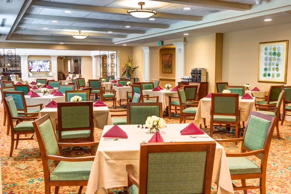 A spacious dining room with multiple tables covered in beige tablecloths, each set with folded burgundy napkins and small floral centerpieces. The chairs have wooden frames with green upholstered seats and backs. The room features warm lighting, decorative wall art, and a coffee station in the background.