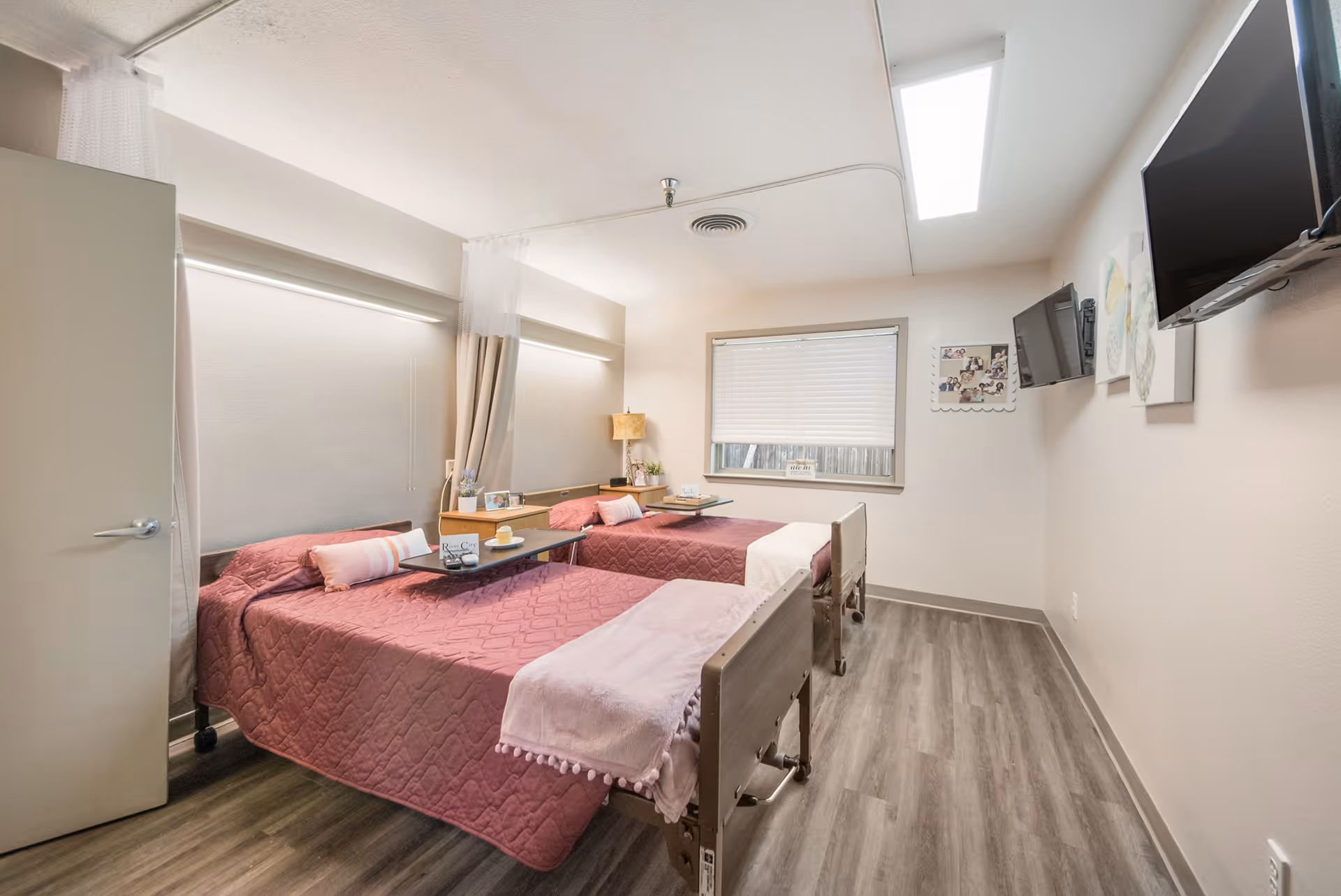 A bright shared resident bedroom with two hospital-style beds covered in pink quilts, overbed tables, wall-mounted TVs, and a window.