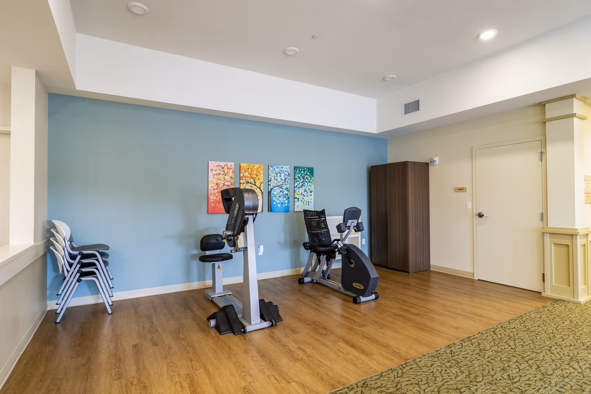 A small exercise room with two stationary exercise bikes placed on a wooden floor. The back wall is painted blue and decorated with four colorful paintings of trees in different seasons. To the left, there is a stack of white and gray chairs. On the right side, there is a closed door and a tall wooden cabinet. The ceiling has recessed lighting.