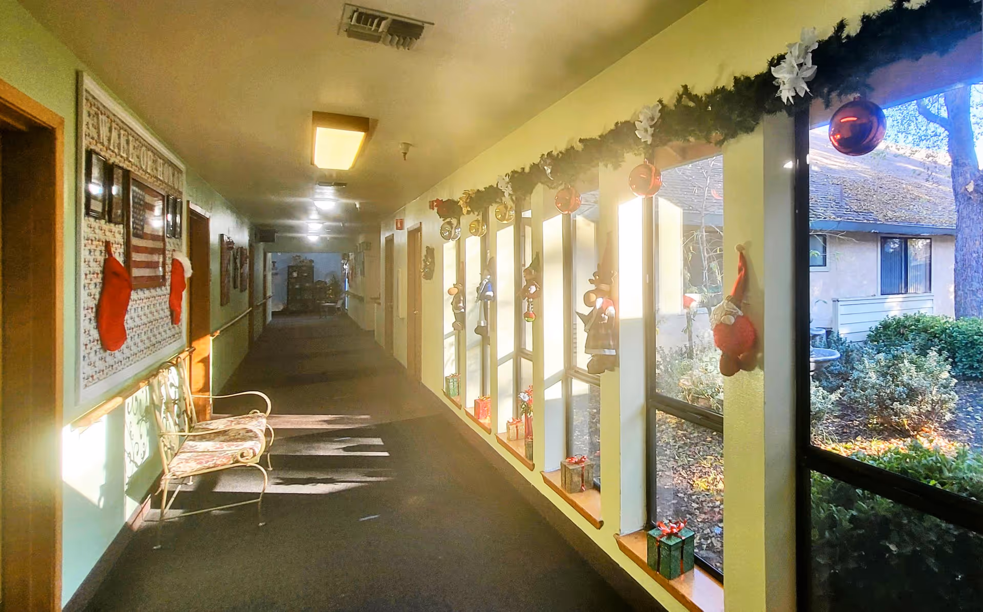 A decorated hallway in Woodland Gardens Senior Living with Christmas stockings hanging on the wall, a bench with floral cushions, and windows adorned with garlands, ornaments, and small wrapped gift boxes. Outside the windows, bushes and a building roof are visible.