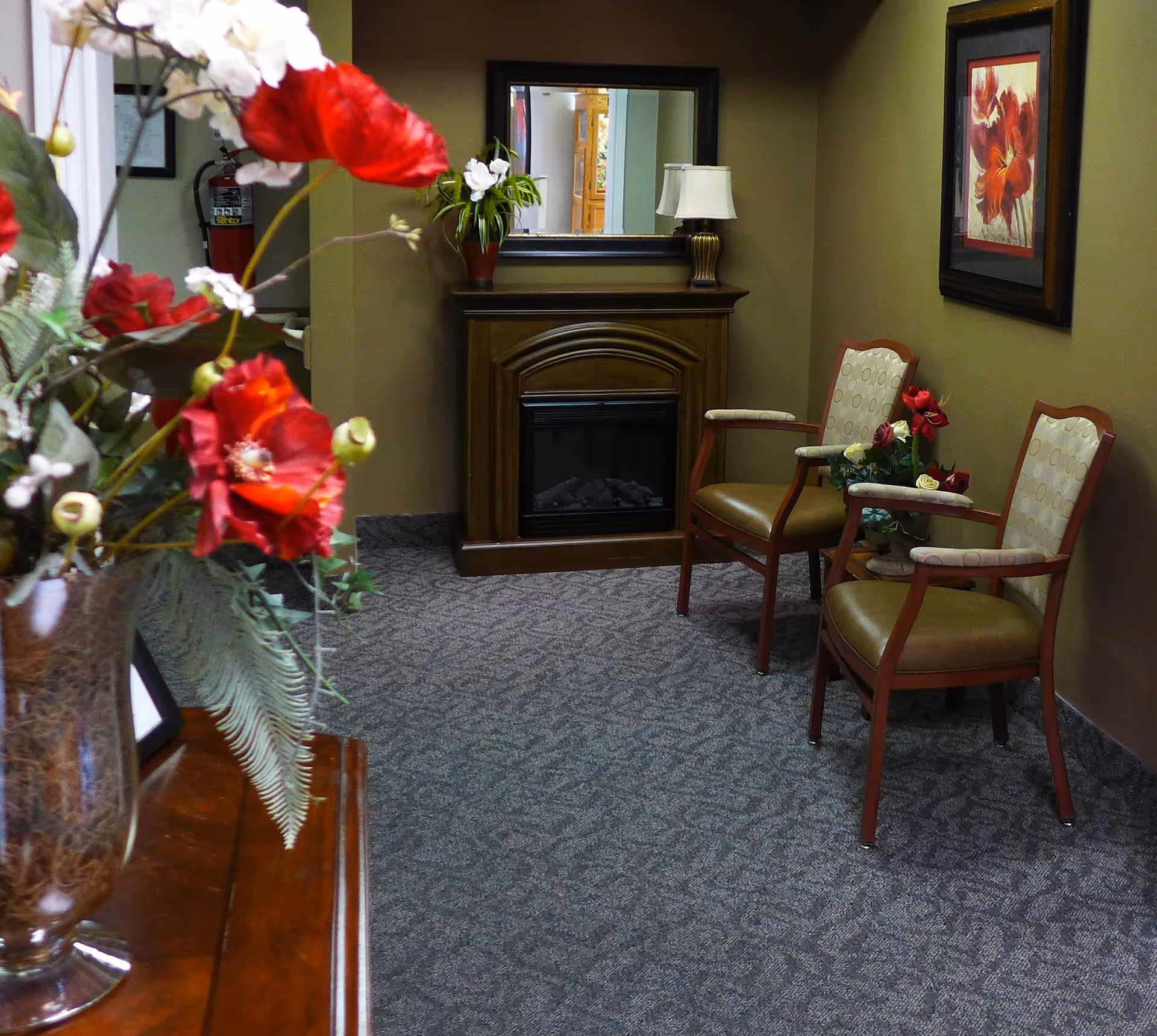 A cozy interior hallway area with two cushioned chairs, a small table with a flower arrangement, a wooden electric fireplace with a lamp and plant on top, a large mirror above the fireplace, and a framed floral painting on the wall. A vase with red and white flowers is partially visible in the foreground.