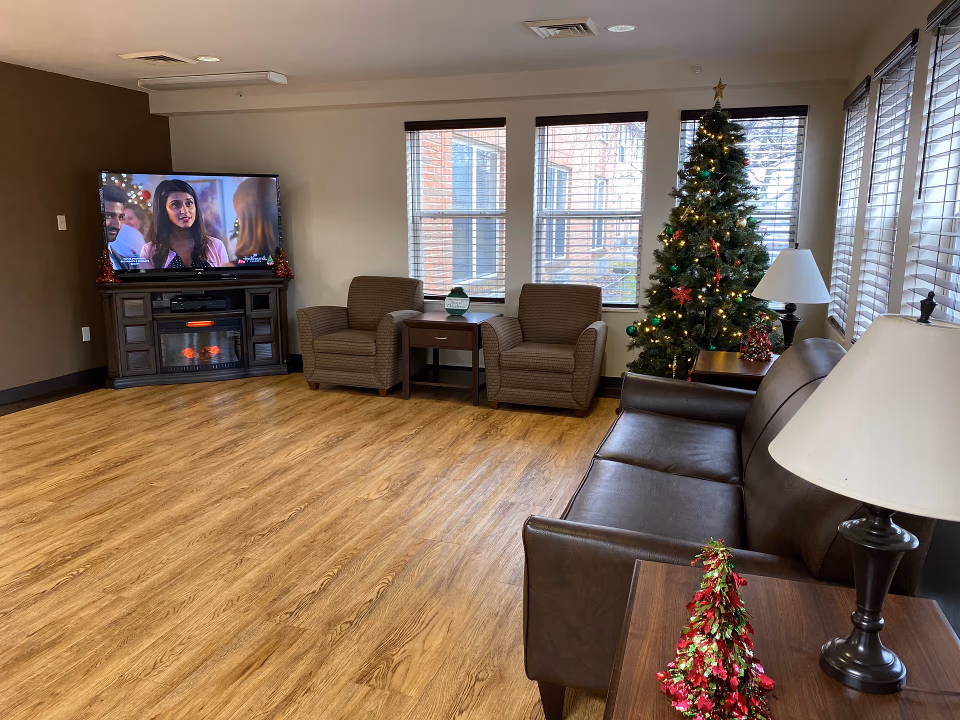 A cozy living room with a brown leather sofa, two brown armchairs, a wooden side table with a lamp and small Christmas tree decoration, a decorated Christmas tree near the windows, and a TV on a stand with an electric fireplace below it. The room has wood flooring and several windows with blinds.