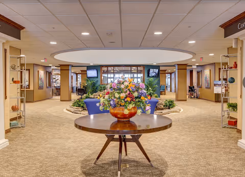 Spacious and well-lit interior common area of Huntley Springs Retirement Resort featuring a round wooden table with a large floral arrangement in the center. The room has beige carpeting, decorative plants, and seating areas with blue chairs. The ceiling has recessed lighting and a large circular light fixture. Walls are adorned with artwork and there are multiple doorways leading to other parts of the facility.