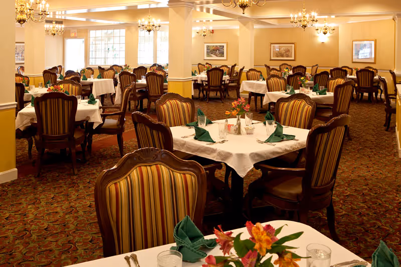 A spacious dining room with multiple tables covered in white tablecloths, each set with green folded napkins, glasses, and floral centerpieces. The room features striped upholstered chairs, chandeliers hanging from the ceiling, framed artwork on the walls, and large windows letting in natural light.