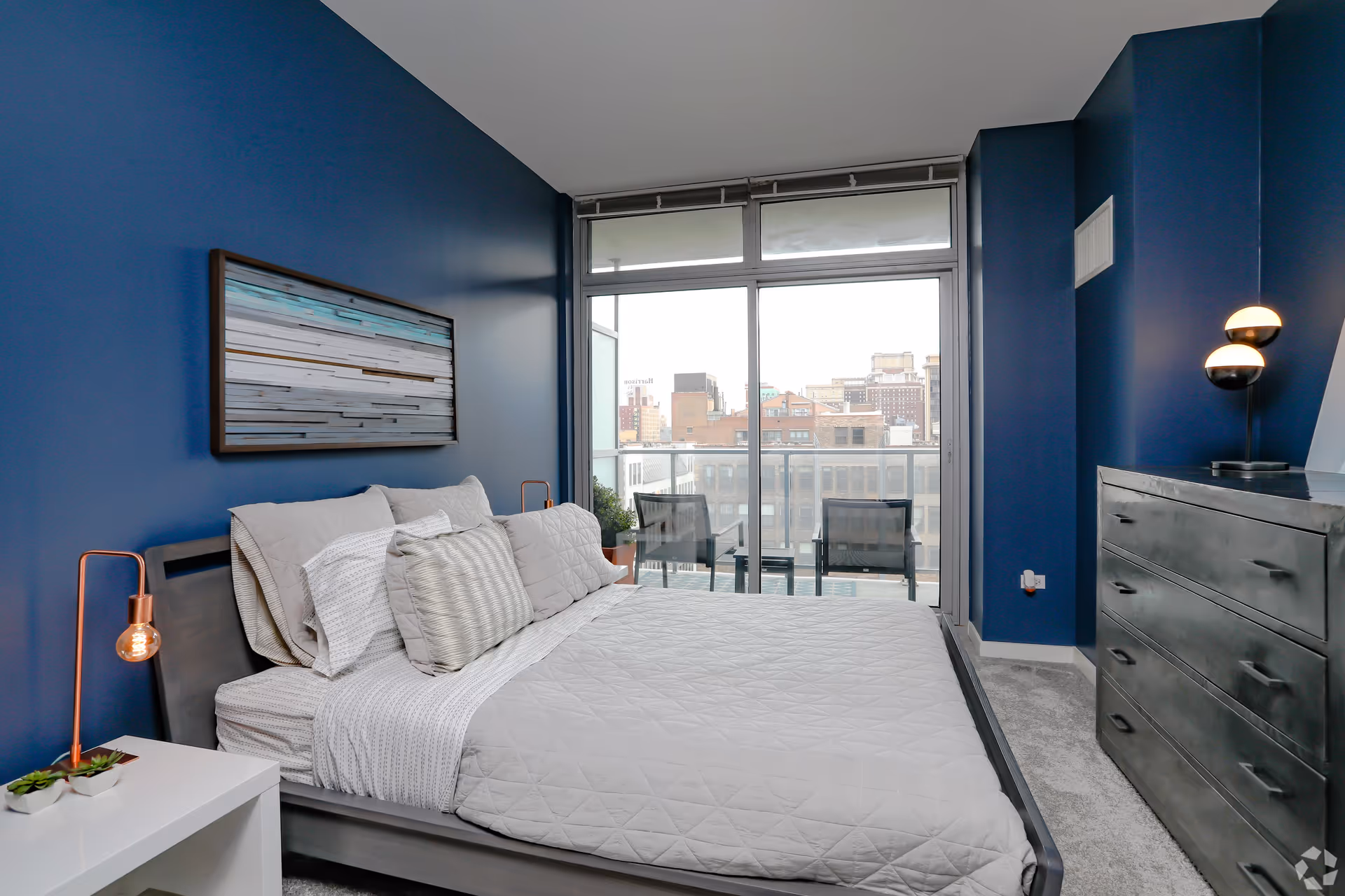 A modern bedroom with dark blue walls, a large bed with white and gray bedding, a white nightstand with a small plant and a copper lamp, a dark gray dresser with a modern lamp on top, and a large glass sliding door leading to a balcony with two chairs and a city view.