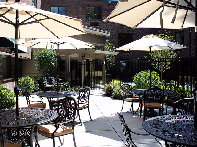 Outdoor patio area with several round metal tables and chairs with cushions, shaded by large beige umbrellas. The patio is surrounded by greenery and bushes, with a brick building in the background.