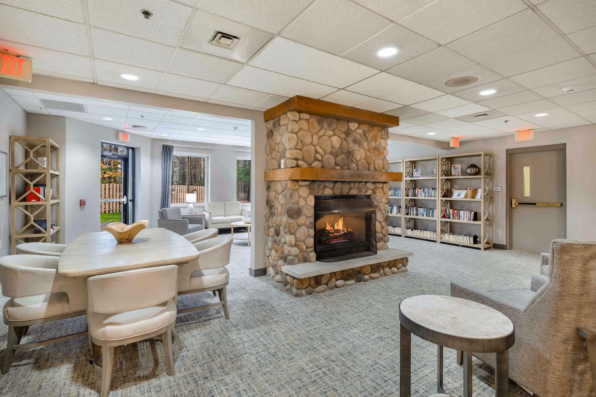 A cozy senior living community common area featuring a stone fireplace with a fire burning, surrounded by comfortable seating including armchairs and a dining table with chairs. Bookshelves filled with books line the walls, and large windows let in natural light. The room has a carpeted floor and a drop ceiling with recessed lighting.