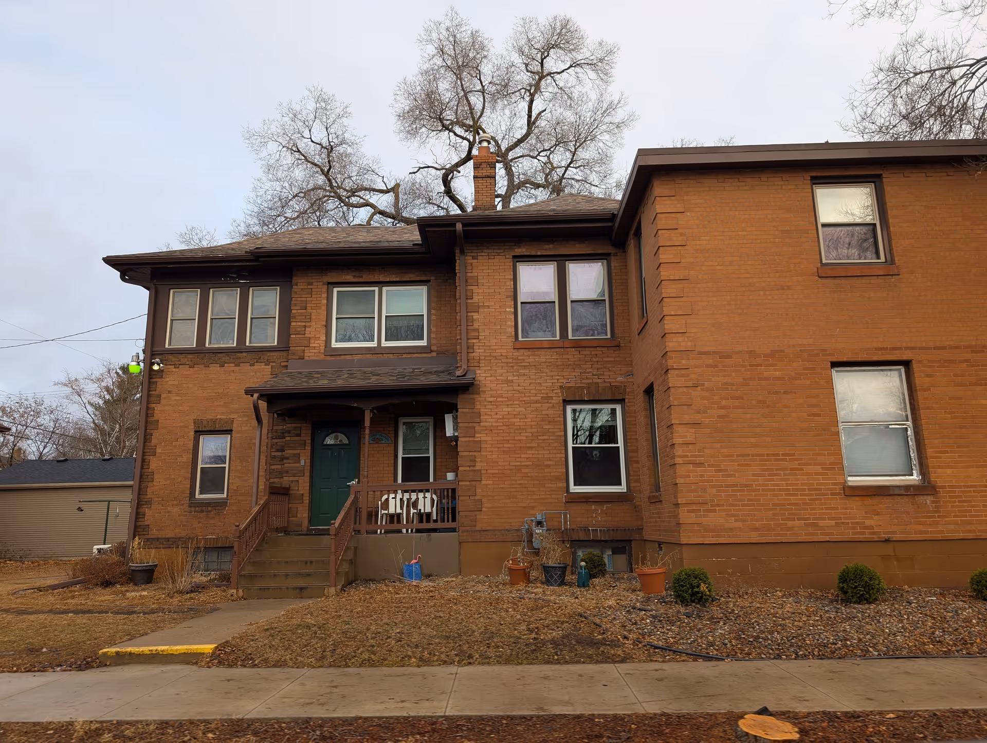 Two-story brick building front with a small porch, green entry door, multiple windows, and leafless trees behind it.