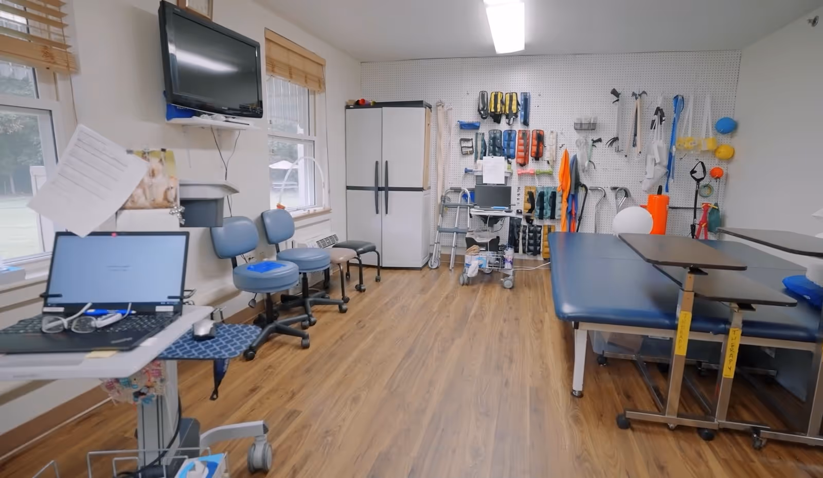 A therapy room with wooden flooring, a blue therapy table, adjustable overbed tables, and a pegboard wall holding various therapy tools and equipment. There are three blue chairs lined up against the wall under two windows, a white refrigerator, a laptop on a mobile workstation, and a wall-mounted TV.