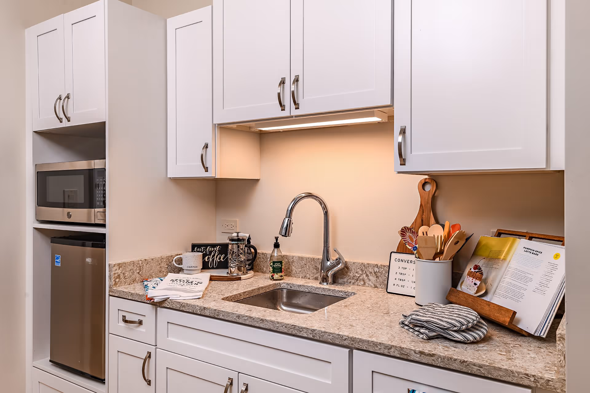 A modern kitchen area with white cabinets and a beige marble countertop. The countertop features a stainless steel sink with a curved faucet, a French press coffee maker, a coffee mug, a soap dispenser, kitchen utensils in a white holder, a wooden cutting board, an open cookbook on a stand, and folded kitchen towels. There is a built-in microwave above a small stainless steel refrigerator on the left side.