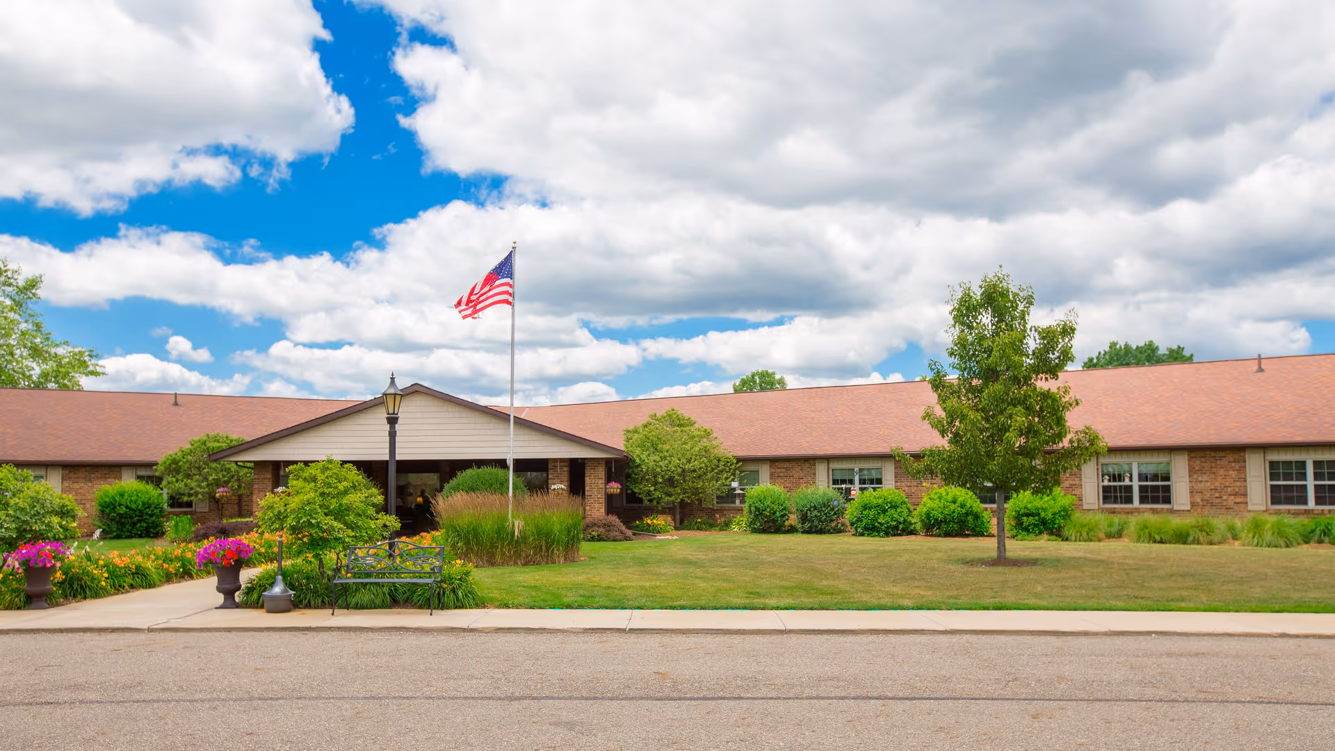 Exterior view of Canterbury Villa of Alliance, a single-story brick building with a brown roof, surrounded by green shrubs, trees, and colorful flower pots. An American flag flies on a flagpole in front of the entrance under a partly cloudy blue sky.