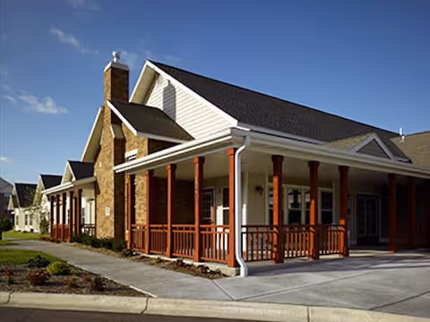 Exterior view of a single-story building with a covered porch supported by wooden columns, brick chimney, and a sloped roof under a clear blue sky.
