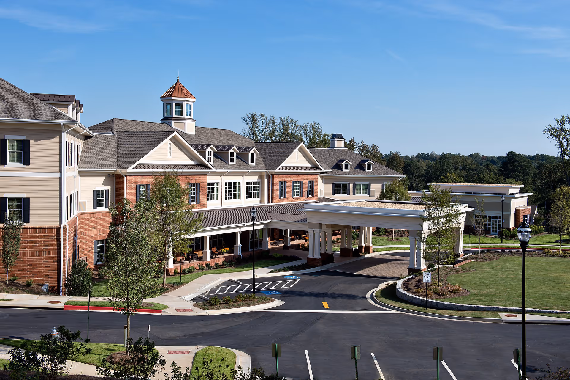 Exterior view of Sterling Estates of East Cobb Retirement Community showing a large, multi-story building with a covered entrance driveway, landscaped grounds, and clear blue sky.