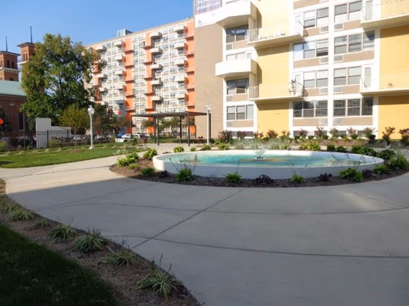 Outdoor courtyard area at City Parc at Pine featuring a circular water fountain surrounded by plants, concrete walkways, and a multi-story residential building in the background under a clear sky.