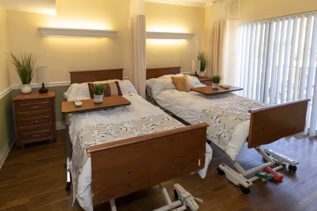 Shared bedroom with two adjustable wooden beds, overbed tables, nightstands, potted plants and vertical blinds.