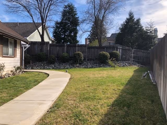 Curved concrete walkway through a grassy fenced backyard with shrubs, trees, and the side of a house visible.