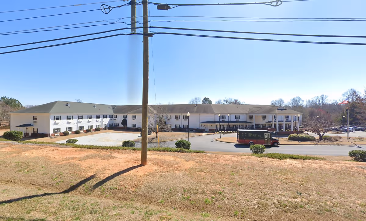 Exterior view of a large two-story senior living facility building with a covered entrance and a shuttle bus parked in front. The building is surrounded by a parking lot, some bushes, and a grassy area with a utility pole and power lines visible in the foreground under a clear blue sky.