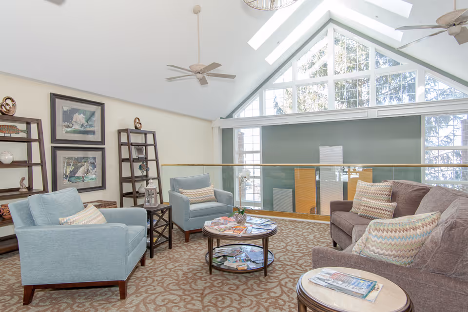 A bright and spacious living room area with high vaulted ceilings and large triangular windows letting in natural light. The room features two light blue armchairs, a brown sofa with patterned cushions, a round coffee table with magazines, and wooden shelving units with decorative items. Ceiling fans hang from the ceiling, and the carpet has a beige and brown pattern.