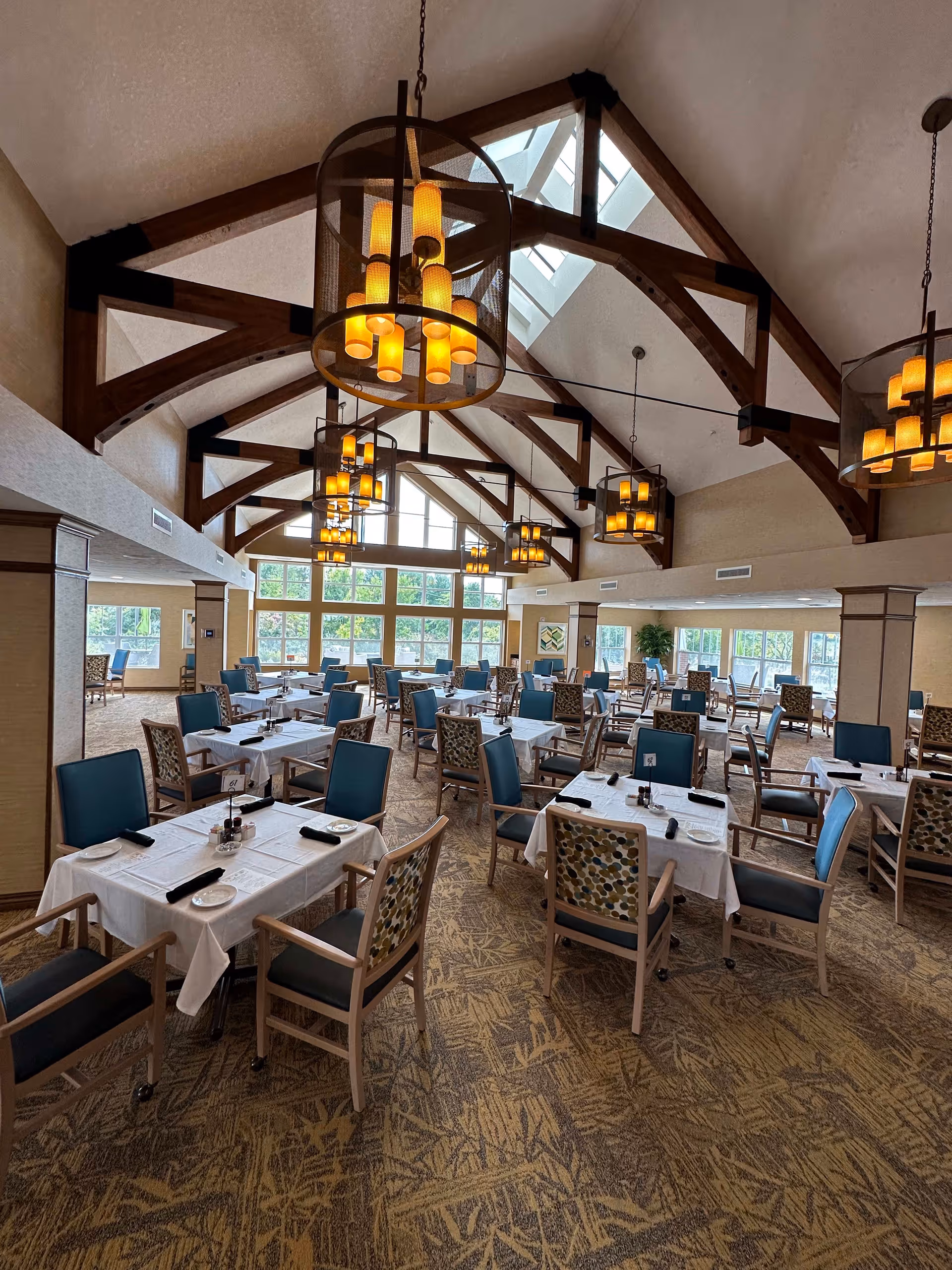 Spacious dining room with multiple tables covered in white tablecloths, each set with plates, napkins, and condiments. The room features high vaulted ceilings with exposed wooden beams and large hanging light fixtures. Large windows along the far wall allow natural light to fill the space, and the carpet has a patterned design.