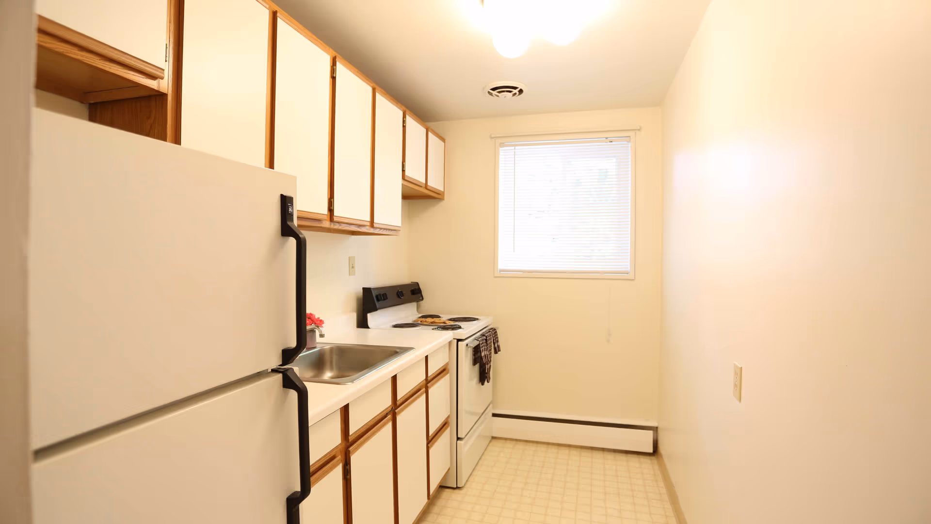 A small kitchen with white cabinets and wooden trim, a white refrigerator, a white stove with oven, a stainless steel sink, and a window with blinds letting in natural light.