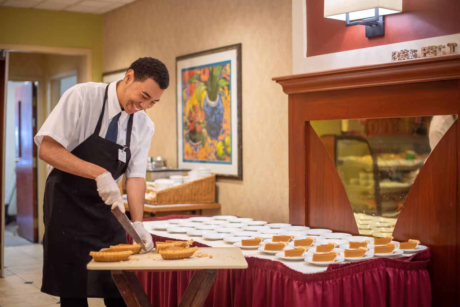 A smiling man wearing a white shirt, black apron, and gloves is slicing pumpkin pies on a table. The table next to him is covered with a red tablecloth and has many slices of pumpkin pie on white plates. The room has beige walls, a framed colorful painting, and a wooden mantel with a wall lamp above it.