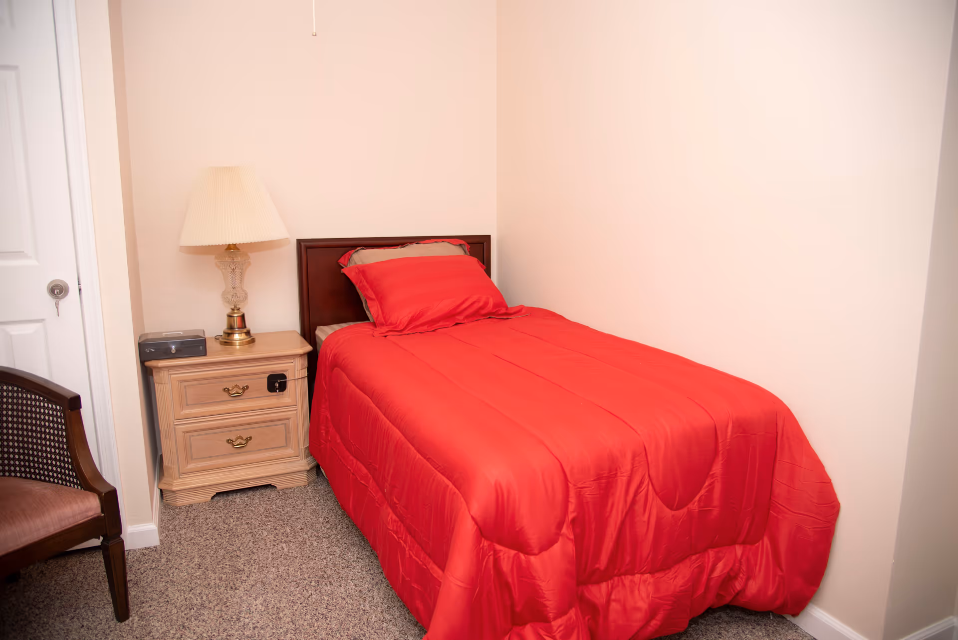Small bedroom with a single bed in a red comforter, a wooden nightstand with a lamp, and a chair.