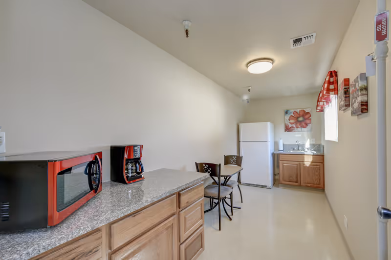 A small kitchen area with wooden cabinets, a granite countertop, a red microwave, and a red coffee maker on the left side. In the background, there is a white refrigerator, a small dining table with two chairs, a sink with a cabinet below, and a window with a red checkered valance. The walls are light-colored and there are decorative flower paintings on the wall near the window.