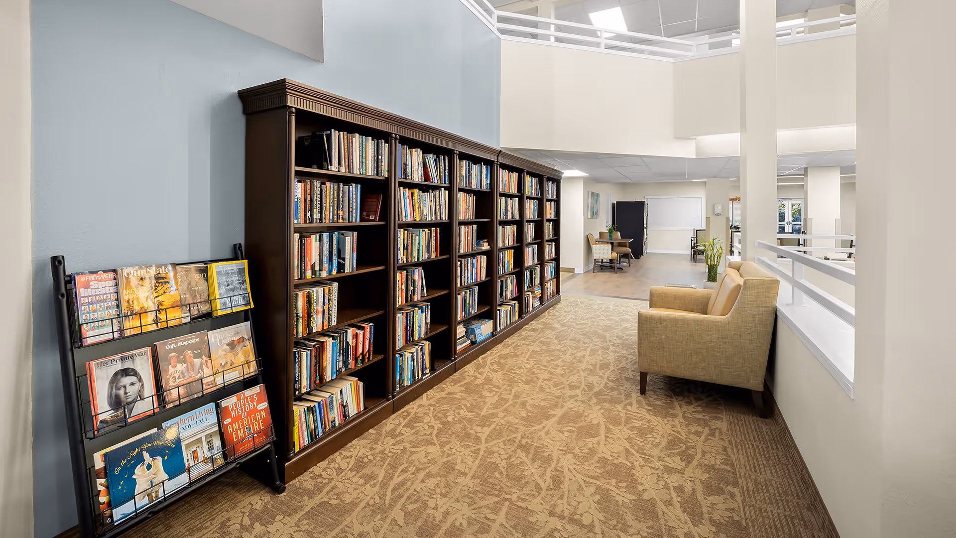 Interior view of a senior living facility library area with a large dark wooden bookshelf filled with books along the left wall, a magazine rack displaying various magazines, a beige armchair on the right side, and an open space leading to a seating area and tables in the background.
