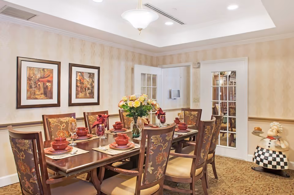 A formal dining room with a wooden table set for eight people. The table is arranged with red bowls, plates, glasses with napkins, and silverware. A vase with yellow and pink flowers is placed in the center. The room has patterned wallpaper, two framed paintings on the wall, a chandelier, and a decorative chef statue holding a tray in the corner. There is a white door with glass panels leading to another room.