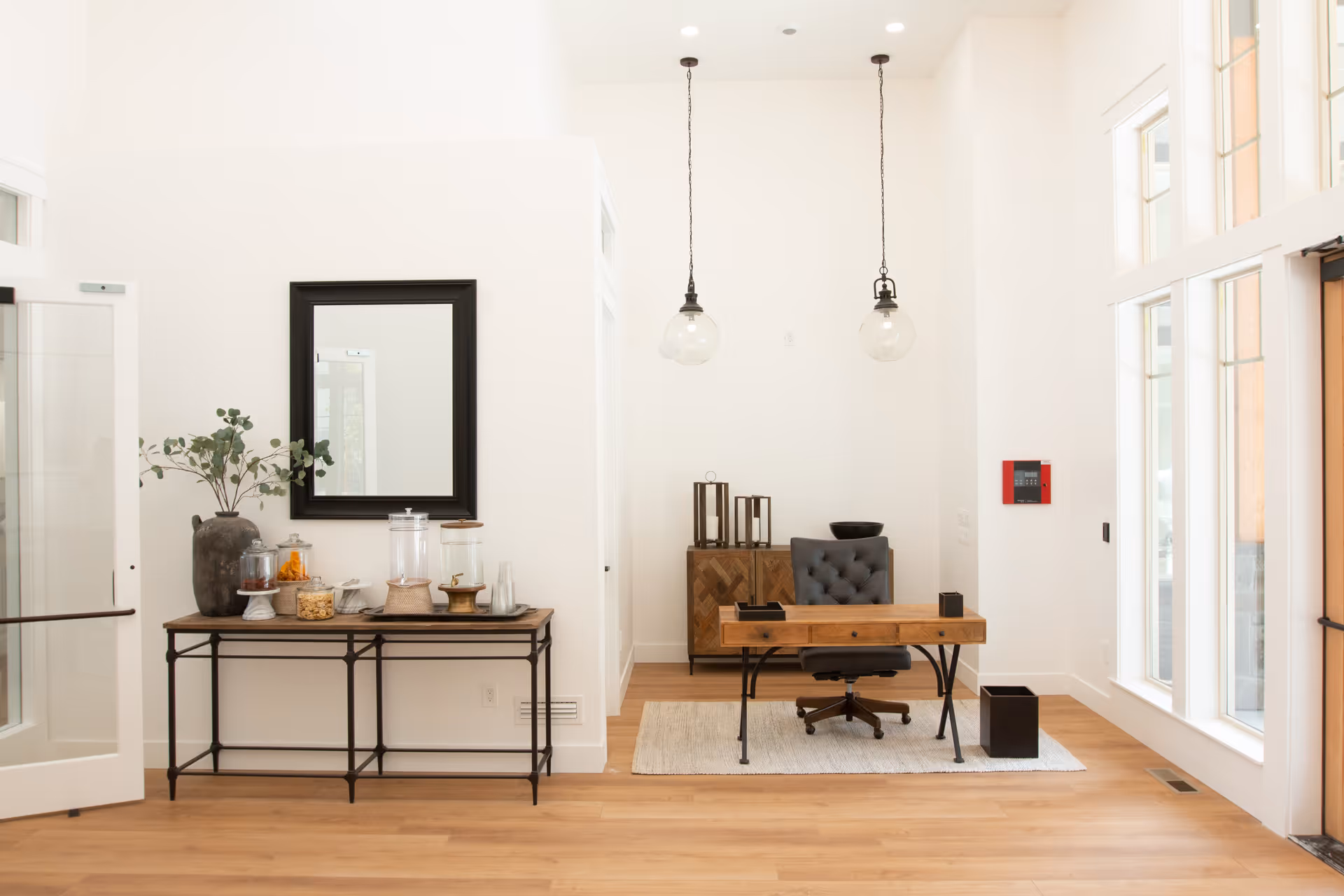 A bright, modern office space with white walls and wooden flooring. There is a wooden desk with black metal legs, a black cushioned office chair, and a small wooden cabinet behind the desk. Two hanging pendant lights with glass globes are suspended from the ceiling. To the left, a console table holds a large vase with greenery, glass containers with snacks, and a mirror above it. Large windows on the right let in natural light.