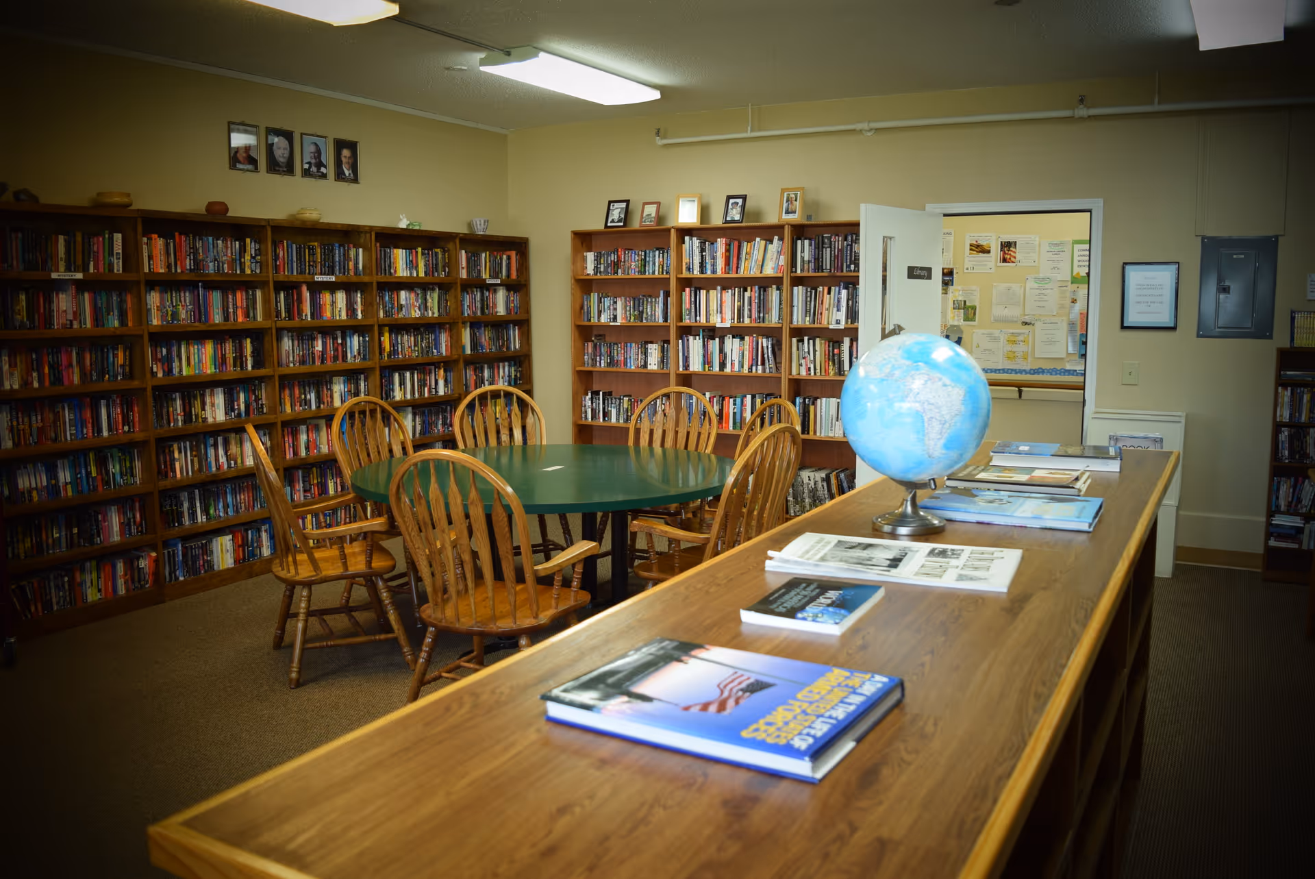 Interior library with bookshelves lining the walls, a round green table surrounded by wooden chairs, and a long counter displaying a globe and books.