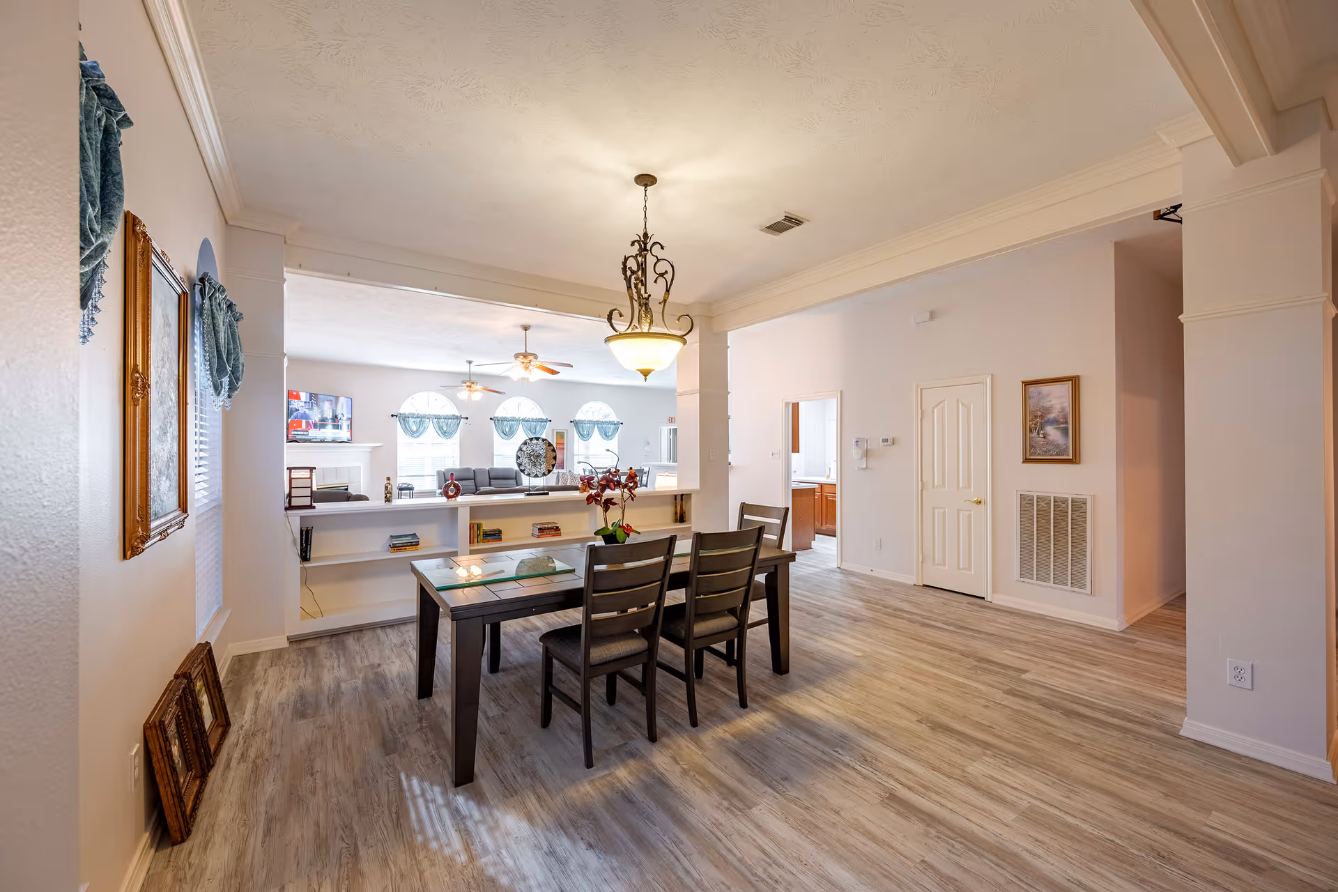 Open dining area with a wooden table and chairs under a chandelier overlooking a bright living room with sofas and arched windows.