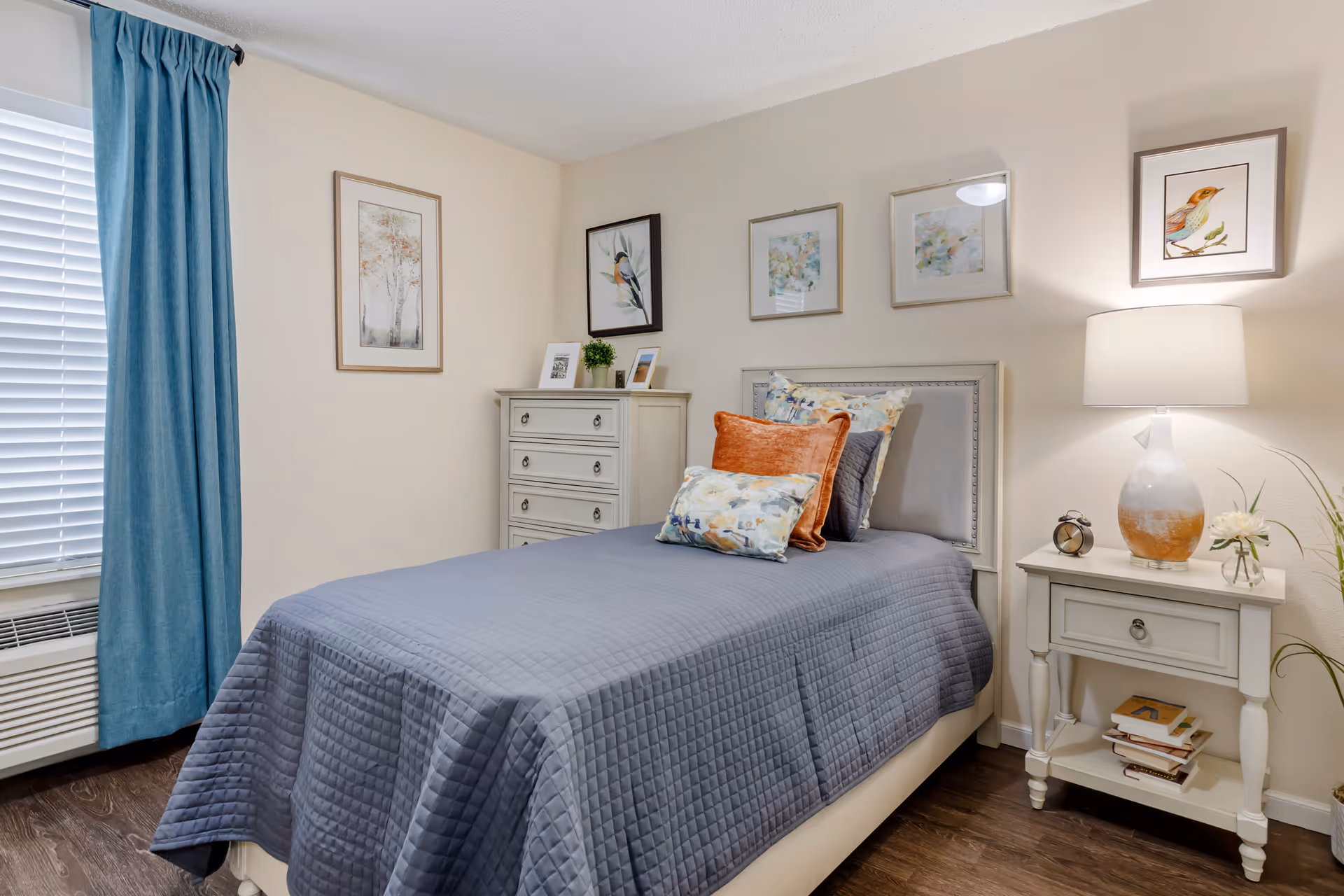 A neatly made single bed with a gray quilted bedspread and multiple decorative pillows in a bedroom. The room features light-colored walls adorned with framed artwork, a white chest of drawers with small framed photos and a plant on top, a white nightstand with a lamp, an alarm clock, and some books. A window with white blinds and blue curtains is visible on the left side.