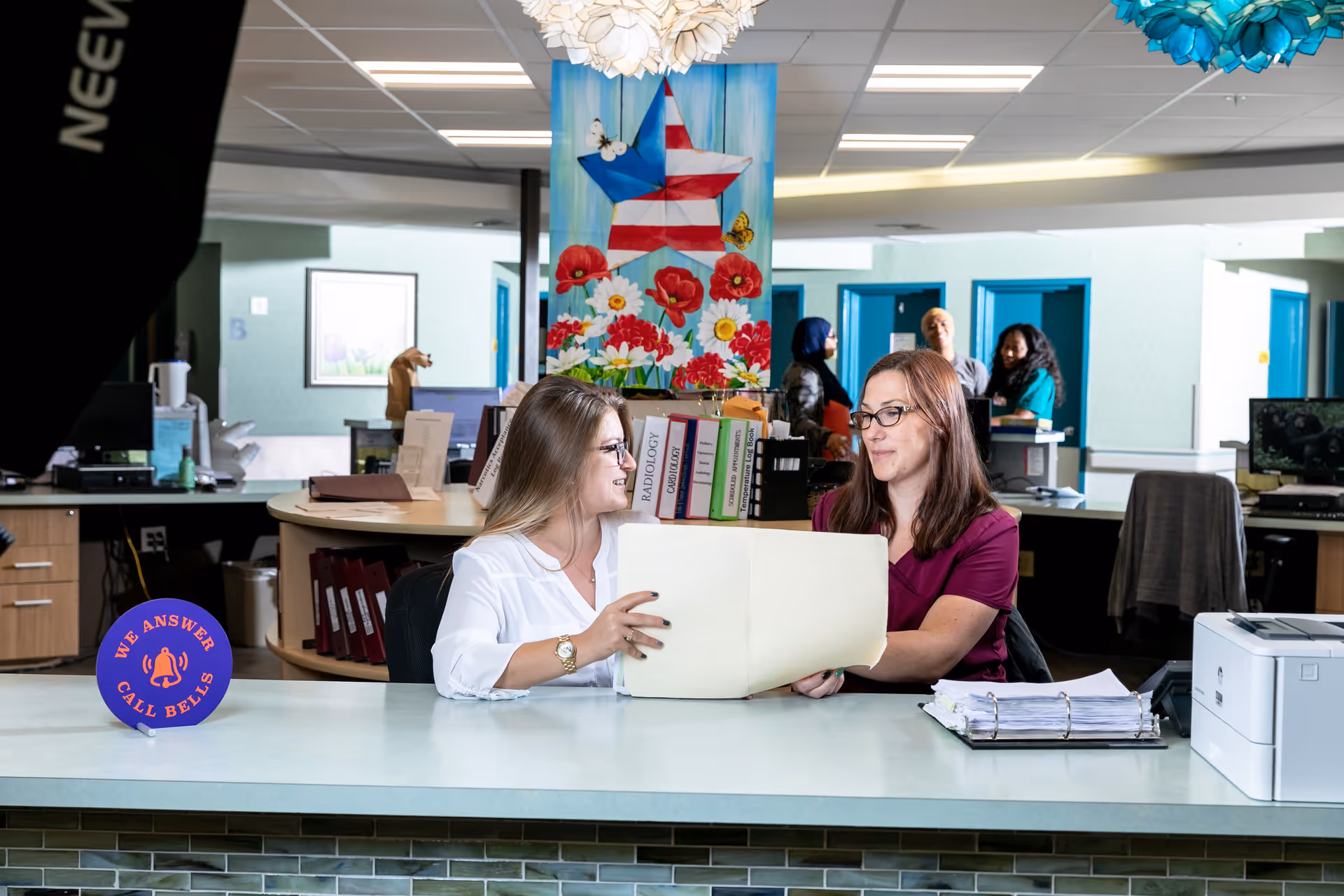 Two women sitting at a reception desk in an office environment, looking at a large folder together. Behind them, there are several people standing and talking near blue doorways. The office has a colorful painting of a star with flowers and butterflies on the wall, and a sign on the desk reads 'We Answer Call Bells.'