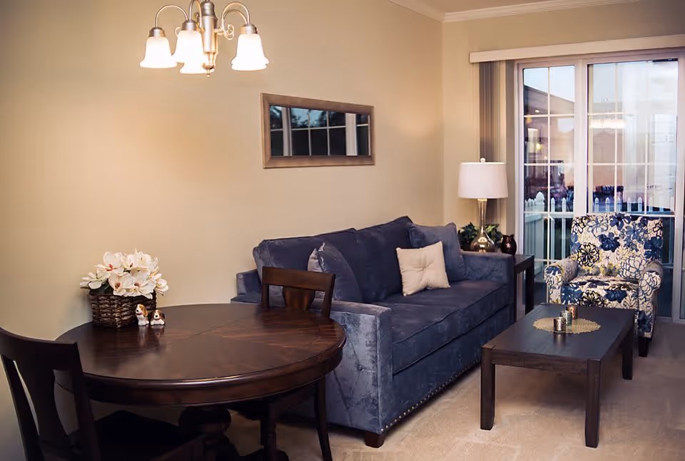 A cozy living room area featuring a dark blue sofa with a beige pillow, a floral patterned armchair, a dark wooden coffee table with decorative items, and a round wooden dining table with two chairs. A basket with white flowers and small figurines sits on the dining table. A lamp with a white shade is on a side table next to the sofa. A window with vertical blinds and a sliding glass door leading to an outdoor area are visible in the background. A wall-mounted mirror and a ceiling light fixture with four lamps are also present.