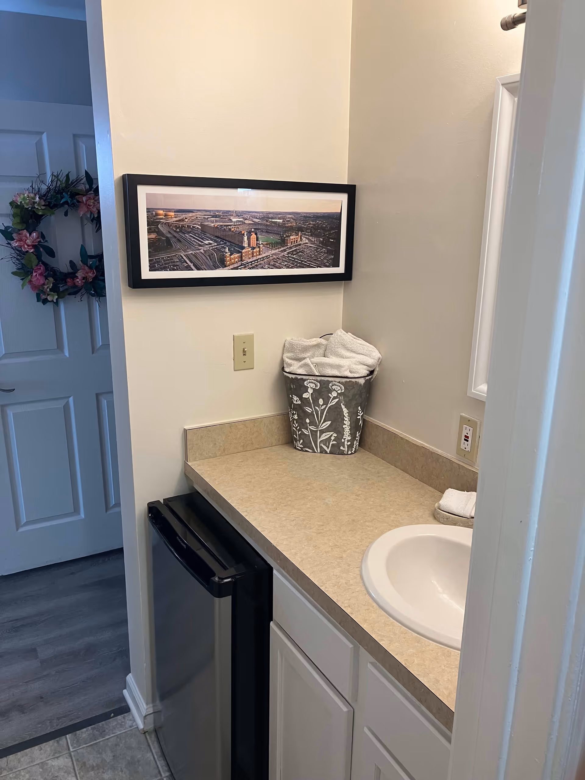 A small bathroom vanity area with a beige countertop, a white sink, and a small black mini-fridge underneath. On the countertop, there is a decorative container holding white towels. A framed panoramic photograph of a cityscape hangs on the wall above the countertop. A door with a floral wreath is partially visible to the left.
