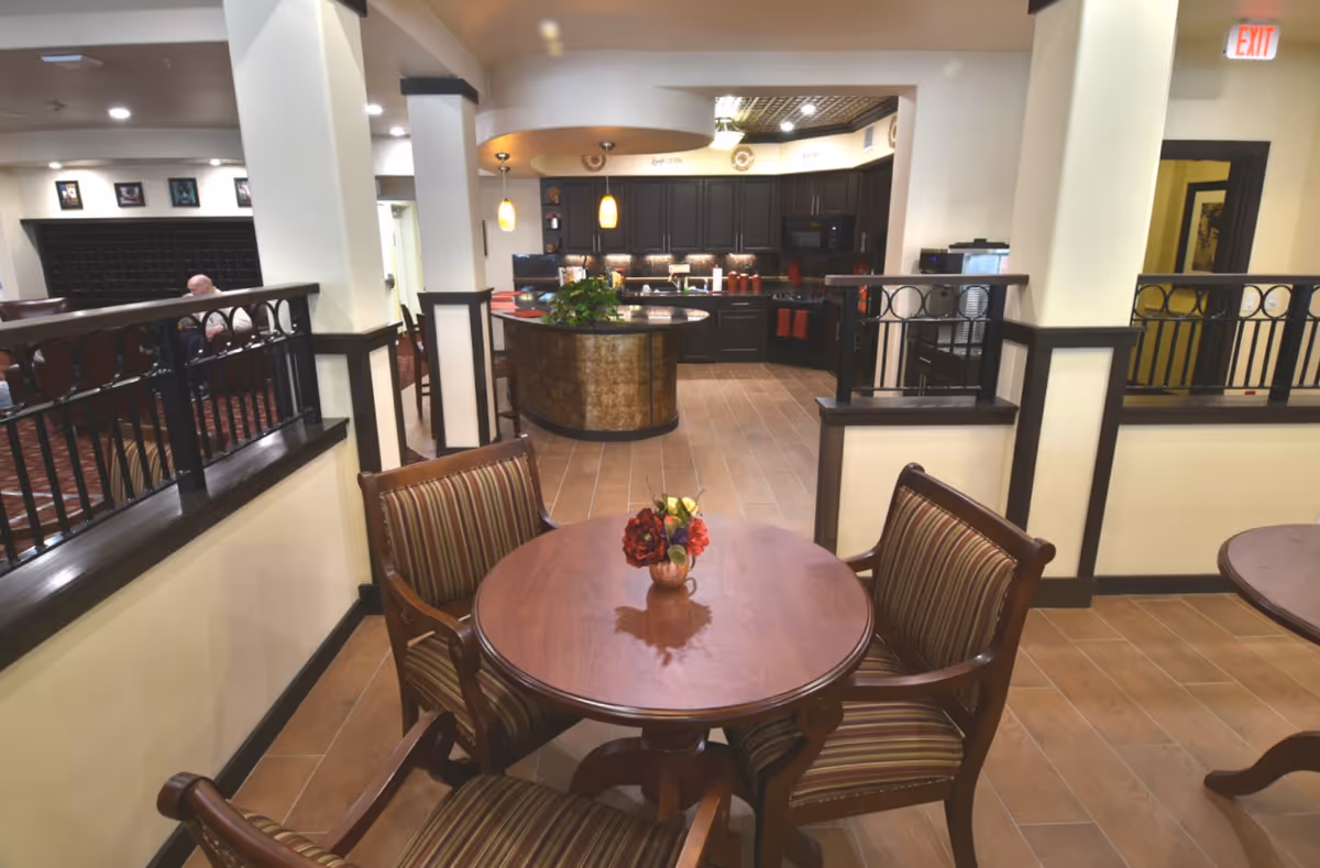 Interior view of a senior living facility showing a cozy seating area with a round wooden table and four striped cushioned chairs. A small vase with red and yellow flowers is placed on the table. In the background, there is a kitchen area with dark cabinets, a curved island with a plant, and pendant lights hanging from the ceiling. The floor is tiled, and the walls are painted in light colors with dark wood trim. A man is seated in the distant background near a wall with mailboxes.
