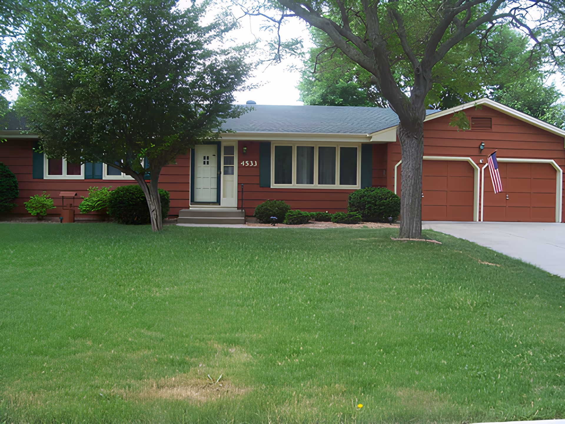 Single-story red house with an attached two-car garage, front lawn, trees, and an American flag.