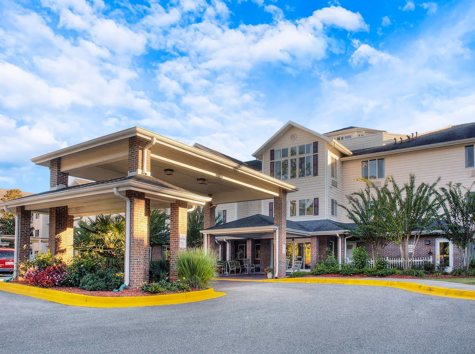 Exterior view of The Woods at Holly Tree Gracious Retirement Living facility showing a covered entrance with brick pillars, landscaped greenery, and a multi-story building under a partly cloudy blue sky.