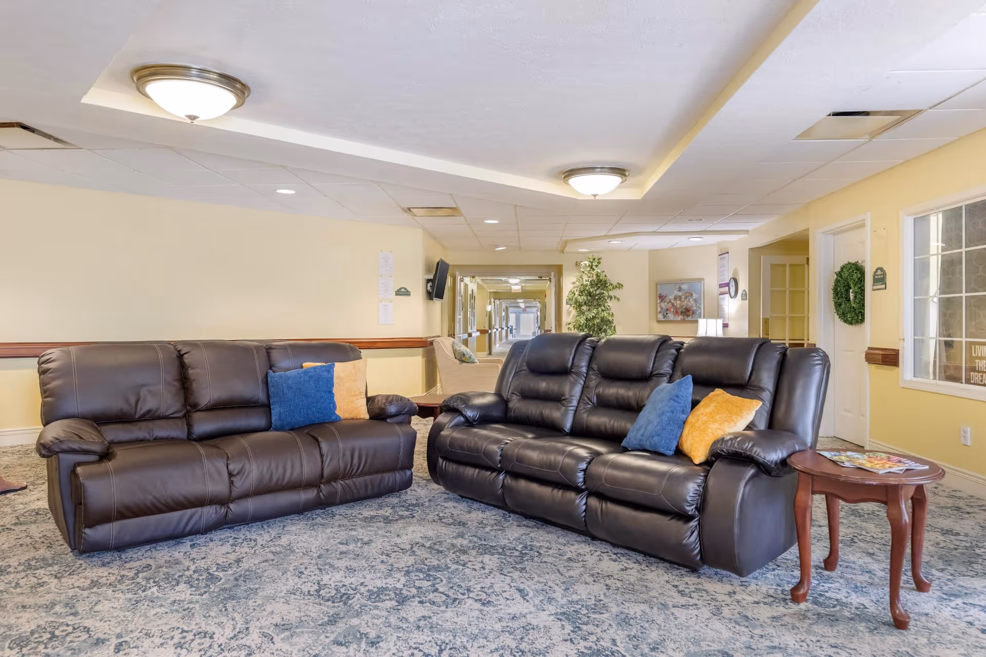 A cozy seating area in a senior living facility featuring two dark brown leather sofas with colorful throw pillows, a small wooden side table with magazines, and a hallway with light yellow walls and ceiling lights in the background.